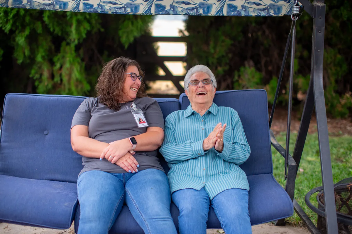 A professional caregiver sitting outdoors with an elderly woman on a porch swing, sharing a joyful moment and providing Home Care in Bend through companionship and emotional support.